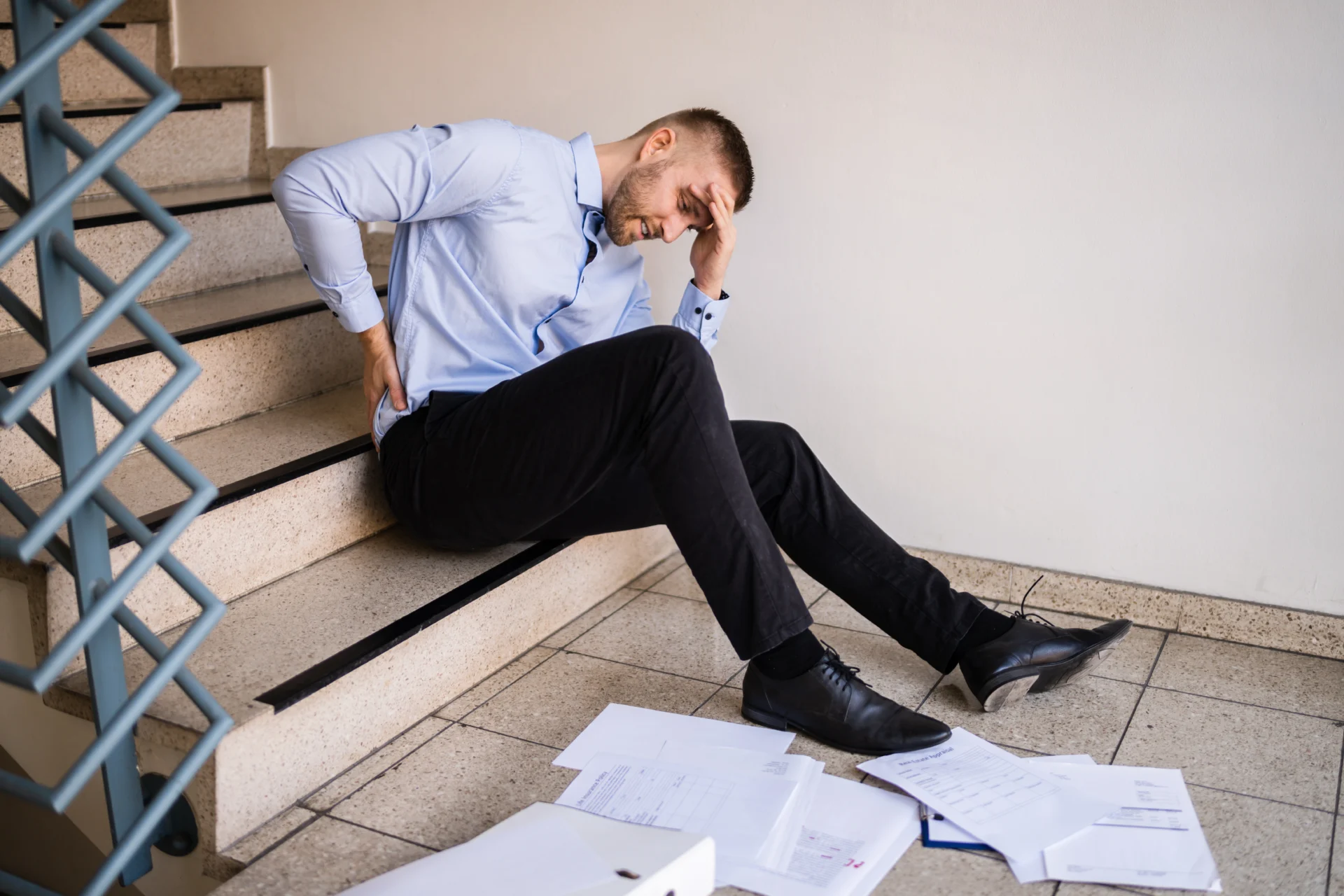 A person holds their back sitting at the base of steps with papers all over the floor