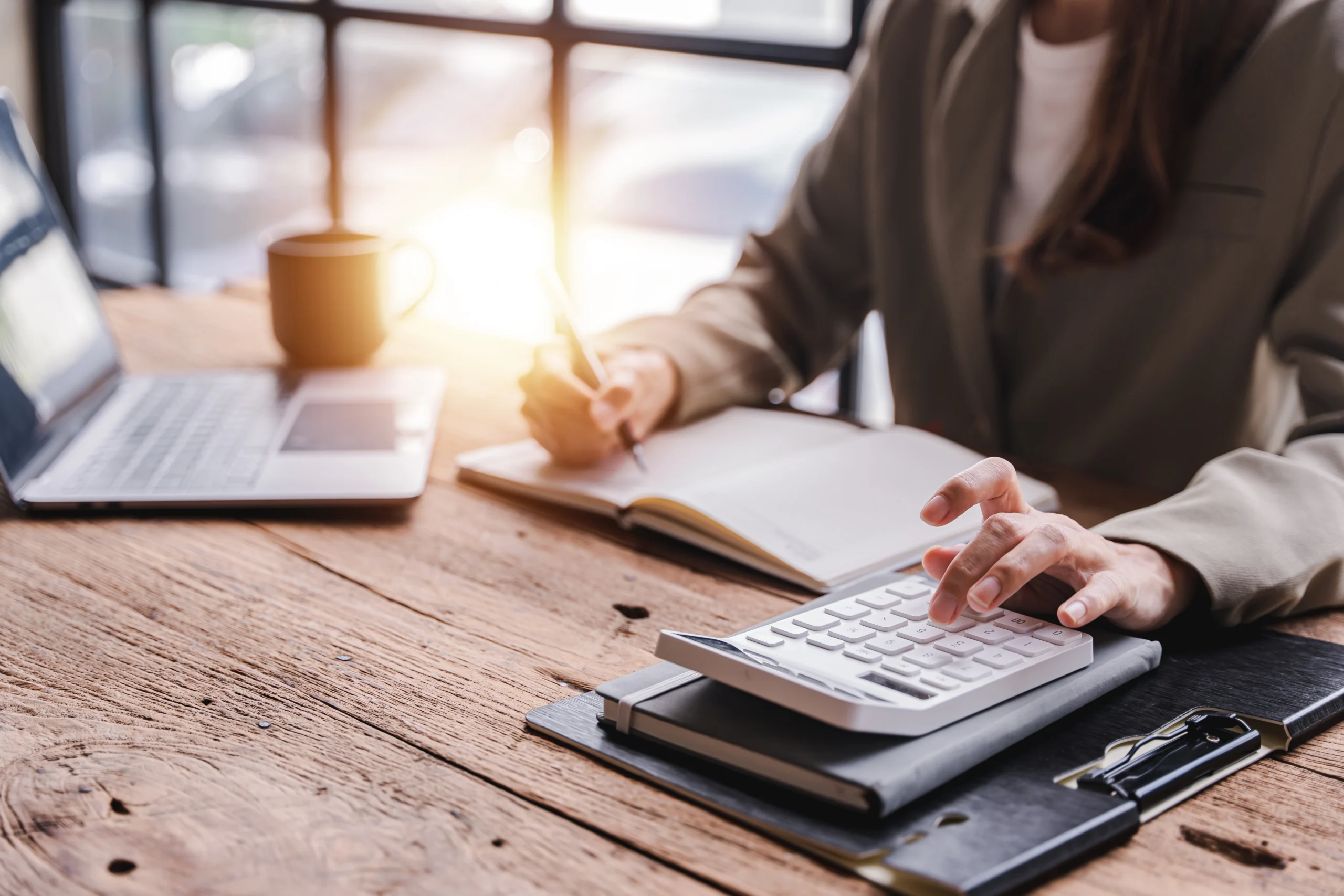A business woman types on a calculator as she writes in a notebook in front of a laptop.